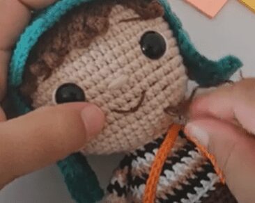 Primer Plano De Unas Manos Que Sujetan Y Cosen La Boca De Una Muñeca Tejida a Crochet Hecha a Mano La Muñeca Tiene Grandes Ojos Negros Cabello Castaño Rizado Una Amplia Sonrisa Y Lleva Un Sombrero Verde Con Orejeras Y Un Traje a Rayas Marrón Y Blanco