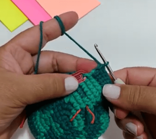 Una Persona Con Esmalte De Uñas De Color Claro Está Tejiendo a Crochet Con Hilo Verde Su Mano Izquierda Sostiene El Hilo Y El Proyecto Mientras Que Con La Derecha Manipula Un Ganchillo En El Fondo Hay Papeles De Colores Brillantes Sobre La Superficie