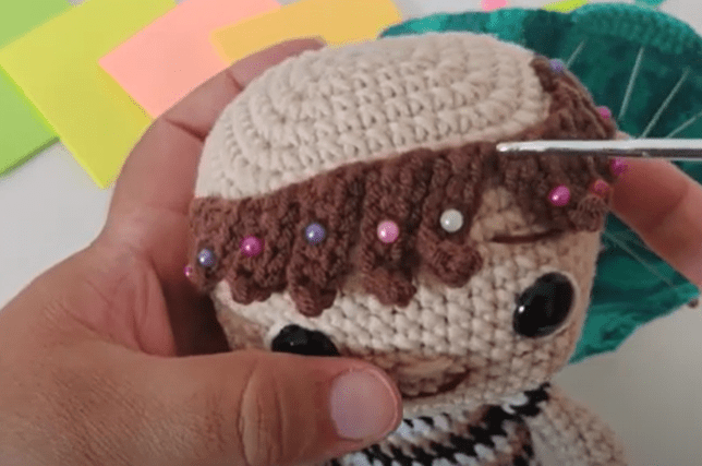 Primer Plano De Unas Manos Trabajando En Una Muñeca Tejida a Crochet Con Pelo Castaño El Pelo Se Fija Con Un Ganchillo Y Se Sujeta Con Alfileres De Colores En El Fondo Hay Cuadrados De Papel De Colores