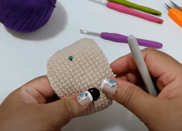 Una Persona Sostiene Con Ambas Manos Una Pequeña Pieza De Tejido a Crochet De Color Beige En El Fondo Sobre La Mesa Hay Varios Ganchos De Crochet Y Una Bola De Hilo Violeta La Persona Tiene Las Uñas Pintadas De Forma Intrincada Con Un Diseño Floral