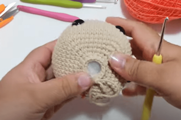 Una Persona Sostiene Un Objeto Tejido a Crochet De Color Beige Claro Con Un Pequeño Agujero En El Centro Utiliza Un Ganchillo Con Mango Amarillo En El Fondo Se Ven Varias Herramientas De Crochet E Hilo Rosa Y Naranja