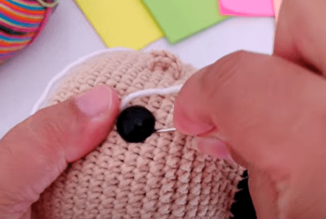 Primer Plano De Unas Manos Cosiendo Una Cuenta Negra Sobre Un Objeto Tejido a Crochet De Color Beige Posiblemente Un Juguete En El Fondo Hay Hojas De Papel De Colores Y Un Objeto De Tela a Rayas