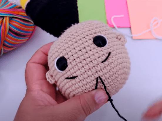 Una Persona Sostiene Una Muñeca Tejida a Crochet Con Cabeza Beige Ojos Negros Y Una Pequeña Sonrisa La Muñeca Tiene El Pelo Negro Hay Una Bola De Lana Multicolor Y Varias Hojas De Papel De Colores En El Fondo