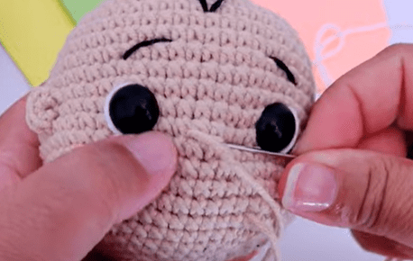 Se Ven Manos Cosiendo Un Juguete De Crochet Beige Con Grandes Ojos Negros Y Cejas Y Cabello Bordados La Pieza Tejida a Crochet Parece Ser La Cabeza De Una Muñeca Los Materiales Coloridos Se Ven Borrosos En El Fondo