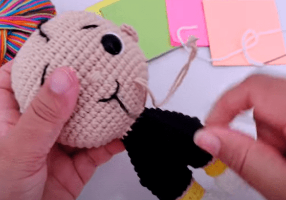 Una Persona Sostiene Una Muñeca Tejida a Crochet Y Termina De Coserle La Cabeza En El Fondo Hay Hilos De Colores Y Algunas Hojas De Papel La Muñeca Tiene Una Cabeza De Color Beige Con Una Cara Divertida Y Viste Un Atuendo Negro