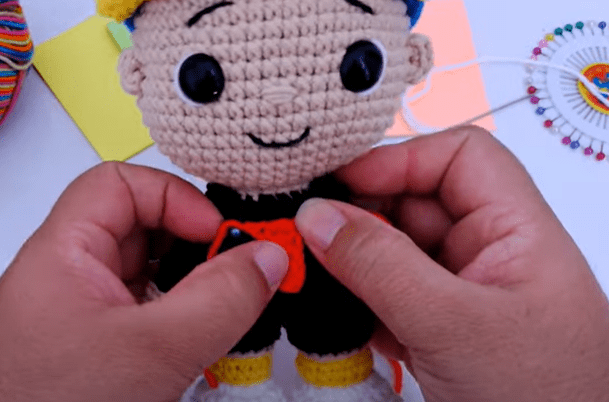 Primer Plano De Unas Manos Que Sostienen Una Muñeca Tejida a Crochet La Muñeca Tiene Una Sonrisa Ojos Negros Grandes Y Lleva Un Sombrero Colorido Las Manos Están En Proceso De Unir Una Pieza Naranja Al Cuerpo De La Muñeca Se Ven Materiales Para Manualidades Y Papel En El Fondo