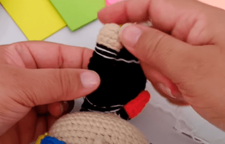 Dos Manos Sostienen Una Pequeña Muñeca Tejida a Crochet En Proceso De Elaboración La Muñeca Tiene Hilo Negro Y Beige Con Algunos Detalles En Rojo Se Ve Papel De Color En El Fondo