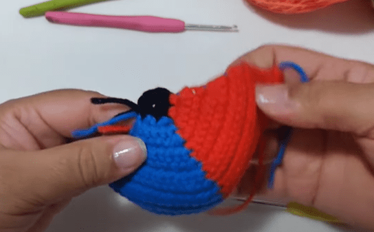 Primer Plano De Unas Manos Que Sostienen Un Objeto Parcialmente Tejido En Crochet De Color Rojo Y Azul En El Fondo Se Ve Un Ganchillo Y Un Poco De Hilo De Colores Azul Rojo Y Naranja La Pieza Tejida Parece Tener La Forma De Una Pequeña Bola O Figura