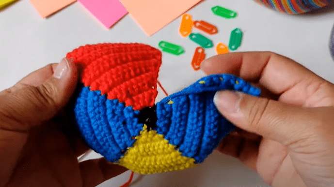 Dos Manos Sostienen Un Objeto De Crochet Colorido Parcialmente Doblado Que Presenta Secciones De Hilo Rojo Azul Amarillo Y Negro Hay Marcadores De Puntos Multicolores Esparcidos Sobre La Mesa En El Fondo Junto Con Notas Adhesivas Y Una Bola De Hilo Rayada