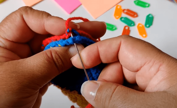 Primer Plano De Unas Manos Tejiendo Una Aguja a Través De Un Hilo De Colores Cosiendo Lo Que Parece Ser Un Proyecto De Crochet O Tejido Marcadores De Puntos De Colores Se Encuentran Dispersos En El Fondo Junto Con Algunas Tarjetas De Notas De Colores Pastel El Enfoque Está En El Trabajo Con La Aguja Y El Hilo