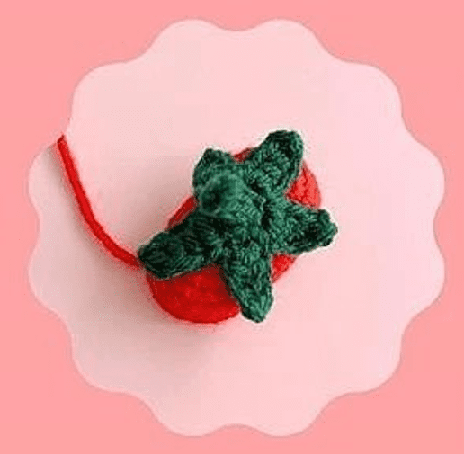 Una Pequeña Fresa Roja Tejida a Crochet Con Hojas Verdes En La Parte Superior Está Centrada Dentro De Un Marco De Borde Festoneado El Fondo Presenta Un Degradado Rosa Suave Que Pasa De Un Rosa Claro En El Centro a Un Rosa Más Intenso En Los Bordes