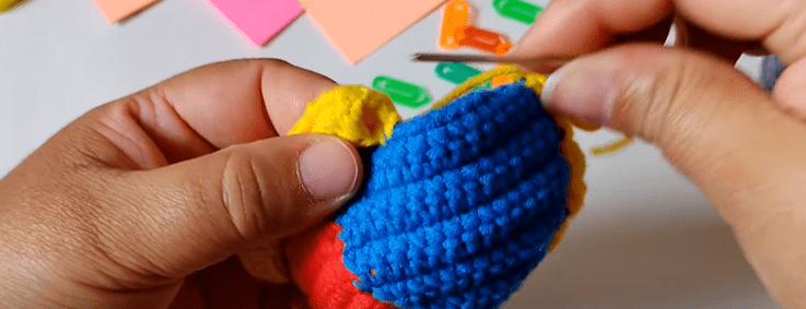 Se Ven Unas Manos Tejiendo Un Objeto Pequeño Y Colorido Con Un Ganchillo El Objeto Está Hecho De Hilo De Color Azul Amarillo Y Rojo Vibrantes En El Fondo Se Ven Parcialmente Sujetapapeles De Colores Y Pequeñas Hojas De Papel