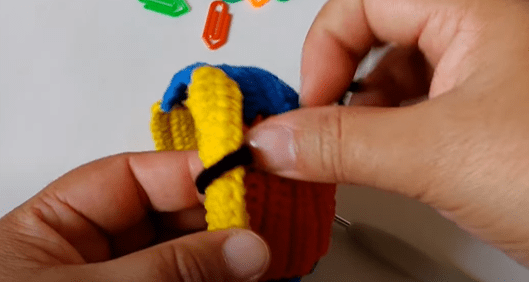 Dos Manos Sostienen Un Pequeño Objeto De Crochet De Colores Principalmente Amarillo Rojo Y Azul El Objeto Parece Ser Una Mochila En Miniatura Varias Etiquetas De Llavero De Colores Brillantes Están Esparcidas Sobre La Mesa Del Fondo