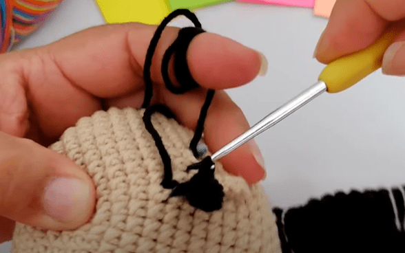 Primer Plano De Unas Manos Tejiendo a Crochet Con Un Hilo Beige Y Negro Una Mano Sostiene La Pieza De Crochet Mientras Que La Otra Manipula Un Ganchillo Con Mango Amarillo Tirando Del Hilo Negro a Través De Un Punto Se Ven Notas Adhesivas De Colores En El Fondo