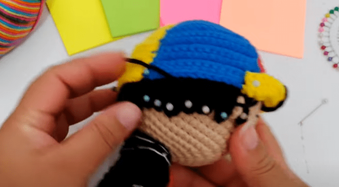 Una Persona Sostiene Un Peluche Tejido a Crochet Que Parece Ser Un Personaje Con Una Gorra Azul Y Amarilla En El Fondo Hay Hojas De Papel De Colores Y Un Alfiletero Circular Con Alfileres