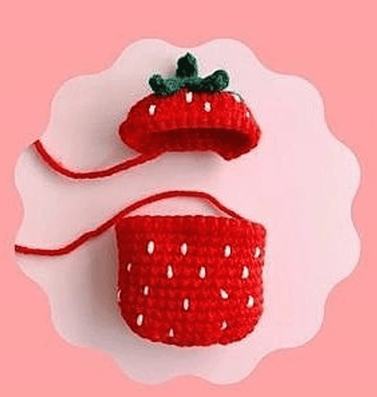 Bolsito De Crochet Con Forma De Fresa Sobre Fondo Rosa La Bolsa Es Roja Con Puntos Blancos En Forma De Semillas Y Tiene Una Hoja De Crochet Verde En La Parte Superior La Tapa De La Bolsa Está Abierta Dejando Al Descubierto Su Interior