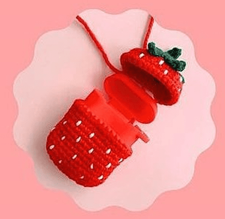 Una Pequeña Bolsa De Crochet Roja Con Forma De Fresa Se Muestra Sobre Un Fondo Rosa Con Bordes Ondulados La Bolsa Tiene Motas Blancas Y Una Parte Superior De Hojas Verdes Su Tapa Está Abierta Revelando Un Recipiente Rojo En Su Interior Se Adjunta Una Cuerda Roja Para Transportarlo