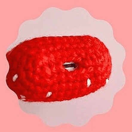 Una Imagen De Primer Plano De Una Fresa Roja Y Blanca Tejida a Mano Con Pequeñas Semillas Blancas Sobre Un Fondo Rosa Con Un Borde Festoneado La Fresa Está Estilizada Y Parece Ser Parte De Un Proyecto Más Grande De Crochet O Amigurumi