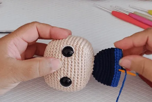 Primer plano de manos tejiendo a crochet la cabeza y la camisa de un amigurumi de Don Ramón.