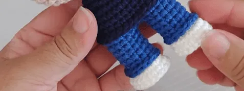 Primer plano de unas manos sosteniendo un amigurumi de Don Ramón tejido a crochet, mostrando pantalones azules y zapatos blancos.