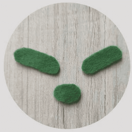 Una Imagen Circular Muestra Un Patrón Simple Parecido a Una Cara Creado Usando Tres Formas Parecidas a Fieltro Verde Sobre Un Fondo De Madera Dos Formas Ovaladas Aparecen Como ojos En La Parte Superior Y Una Forma Ovalada Más Pequeña Sirve Como boca O nariz En El Centro