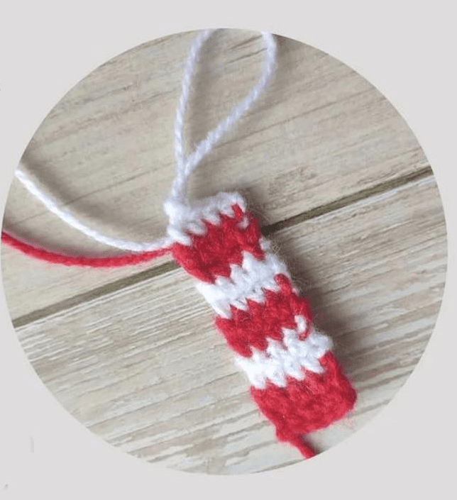 Primer Plano De Una Pieza De Crochet Roja Y Blanca Que Se Asemeja a Un Bastón De Caramelo a Rayas Con Extremos Sueltos De Hilo Que Se Extienden Desde La Parte Superior La Pieza Se Coloca Sobre Una Superficie De Madera Clara