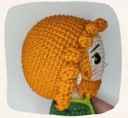 La Imagen Muestra Una Vista Lateral De Un Muñeco Amigurumi Con Pelo Rizado De Color Naranja Y Barba El Duende De La Fortuna Tiene Grandes Ojos Blancos Y Negros Y Viste Un Traje Verde El Fondo Es Completamente Blanco