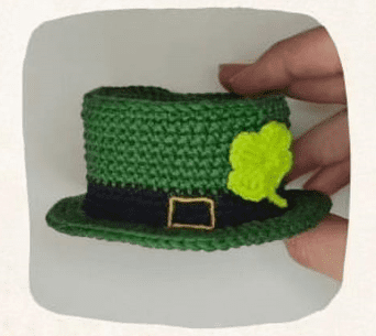 Un Sombrero De Copa Verde De Ganchillo Está Decorado Con Un Trébol Amarillo Y Una Banda Negra Con Una Hebilla Dorada El Sombrero Parecido a Algo Que Podría Usar Un Duende De La Fortuna Está Sostenido Por La Mano De Una Persona En El Lado Derecho De La Imagen