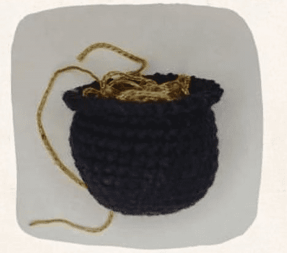 Una Pequeña Olla Tejida De Color Negro Se Asienta Sobre Una Superficie Blanca Contiene Hilo De Color Dorado Entrelazado Que Se Extiende Fuera De La Olla Como Si Esperara Que Un Duende De La Fortuna Le Diera Vida Con La Magia Del Tejer