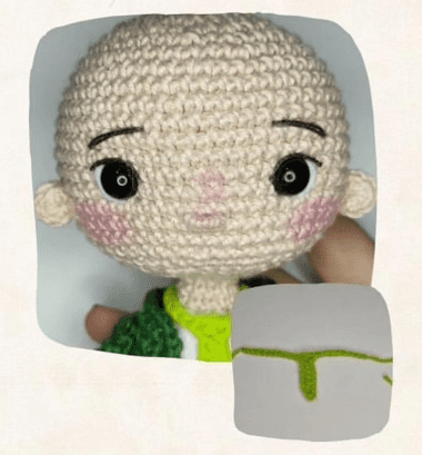 Un Muñeco Amigurumi Tejido a Crochet Con La Cabeza Calva Ojos Negros Y Mejillas Rosadas La Muñeca Lleva Un Traje Verde Claro En La Parte Inferior Derecha De La Imagen Hay Un Bordado Parcial De Un Brote Verde Sobre Un Fondo Blanco Perfecto Para Quienes Buscan Un Patrón Gratuito Para Tejer