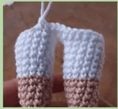 Un Primer Plano De Una Mano Que Sostiene Una Pieza De Crochet En Progreso La Pieza De Crochet Consta De Dos Formas Cilíndricas De Color Azul Claro Unidas En La Parte Superior Con La Parte Inferior En Un Color Marrón Claro Que Recuerda Al Estilo Vibrante De Mirabel Madrigal Parecen Las Etapas Iniciales De Un Par De Pantalones Pequeños