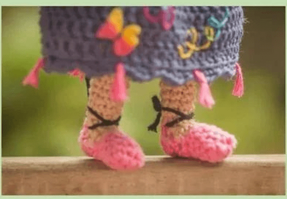 Imagen De Primer Plano De Las Piernas Y Los Pies De Una Muñeca De Crochet La Muñeca Lleva Unos Zapatos De Crochet Rosas Y Un Vestido De Crochet Azul Adornado Con Coloridos Diseños De Mariposas Y Borlas Rosas Que Recuerdan a Mirabel Madrigal El Fondo Está Suavemente Borroso Con Vegetación