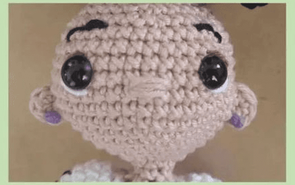 Una Imagen De Primer Plano De La Cabeza De Una Muñeca De Mirabel Madrigal Tejida a Crochet La Muñeca Tiene Grandes Ojos Negros Espesas Cejas Negras Y Una Nariz Pequeña El Fondo Circundante Tiene Un Color Beige Parduzco Claro El Intrincado Trabajo De Crochet Resalta Las Características Detalladas Y Artesanales De La Muñeca