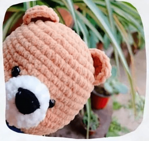 Un Primer Plano De Un Oso Amigurumi Teddy Asomándose Desde El Lado Izquierdo Del Marco El Oso Tiene El Cuerpo Beige La Nariz Negra Y El Hocico Blanco Al Fondo Las Plantas Verdes En Macetas Y La Vegetación Borrosa Sugieren Un Entorno De Jardín Perfecto Para Quienes Buscan Un Patrón Amigurumi Gratis O Tutorial