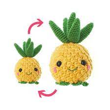 Dos peluches de piña tejidos a crochet con hojas verdes y cuerpo amarillo. Ambas piñas tienen ojos de botón negros, mejillas rosadas y bocas sonrientes. Uno es más grande que el otro, ya que las flechas rojas indican una comparación entre sus tamaños. Perfecto para mostrar técnicas de crochet.