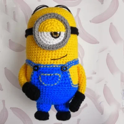 Se muestra un juguete tejido a crochet que se asemeja a un Minion, un personaje de la serie "Mi villano favorito". El juguete tiene un solo ojo, un cuerpo amarillo, un mono azul, guantes negros y zapatos negros. El fondo presenta un patrón de plátanos de color rosa claro sobre un fondo blanco.