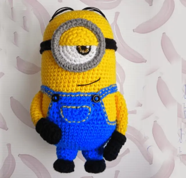 Se Muestra Un Juguete Tejido a Crochet Que Se Asemeja a Un Minion Un Personaje De La Serie mi Villano Favorito El Juguete Tiene Un Solo Ojo Un Cuerpo Amarillo Un Mono Azul Guantes Negros Y Zapatos Negros El Fondo Presenta Un Patrón De Plátanos De Color Rosa Claro Sobre Un Fondo Blanco