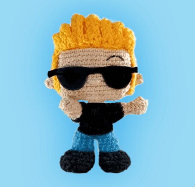 Una Muñeca De Ganchillo Con Cabello Amarillo Brillante Que Lleva Gafas De Sol Negras Camisa Negra Pantalones Azules Y Zapatos Negros El Muñeco Adopta Una Pose Segura Con Una Mano En La Cadera Y La Otra Apuntando Hacia Adelante El Fondo Es De Un Color Azul Claro Sólido