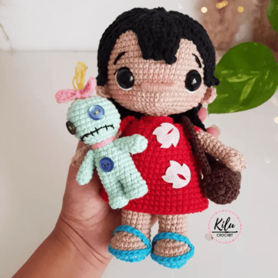 Una mano que sostiene una muñeca de crochet amigurumi con cabello negro, un vestido rojo con estampados blancos y zapatos azules. La muñeca de Lilo va acompañada de una pequeña figura tejida a crochet de color verde con ojos de botón y un lazo rosa en la cabeza. El fondo presenta una planta y el logo "Kilu Crochet".