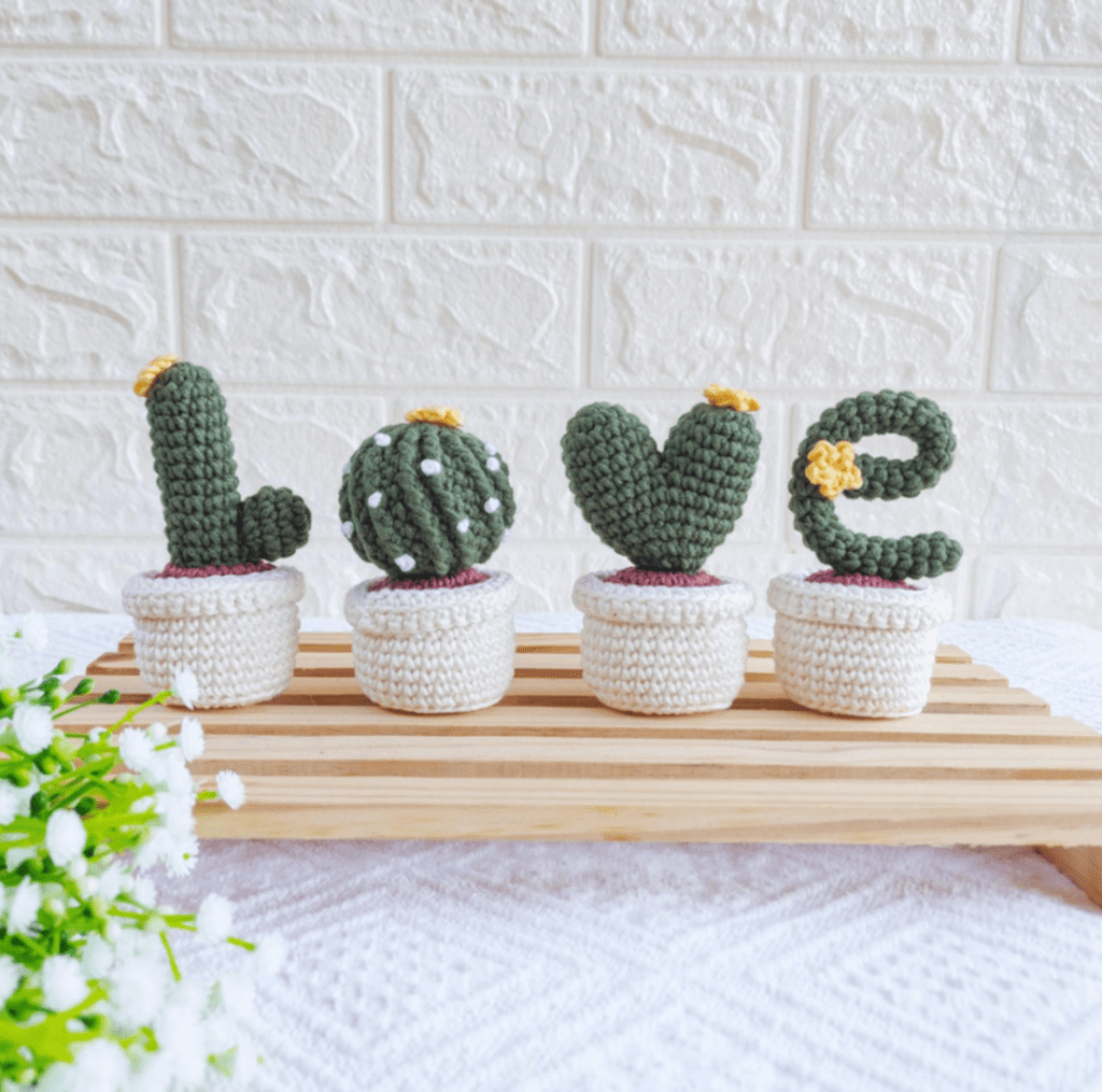 Cuatro Pequeños Cactus De Crochet Cada Uno Con La Forma De Una Letra Que Deletrea amor Se Colocan En Macetas De Crochet Blancas Sobre Una Bandeja De Madera Cada Cactus Amigurumi Tiene Detalles únicos Como Una Pequeña Flor Amarilla Y Manchas Blancas El Fondo Presenta Una Pared De Ladrillo Blanco Texturizado