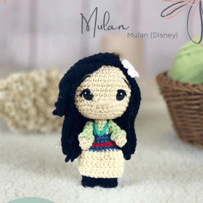 Una muñeca de ganchillo que representa a Mulan de Mulan de Disney. La muñeca tiene el pelo largo y negro, un traje colorido con un top verde, un cinturón azul, una falda beige y una flor blanca en el pelo. El fondo tiene una cesta de crochet y un escenario suavemente difuminado. Texto: "Mulan (Disney)".