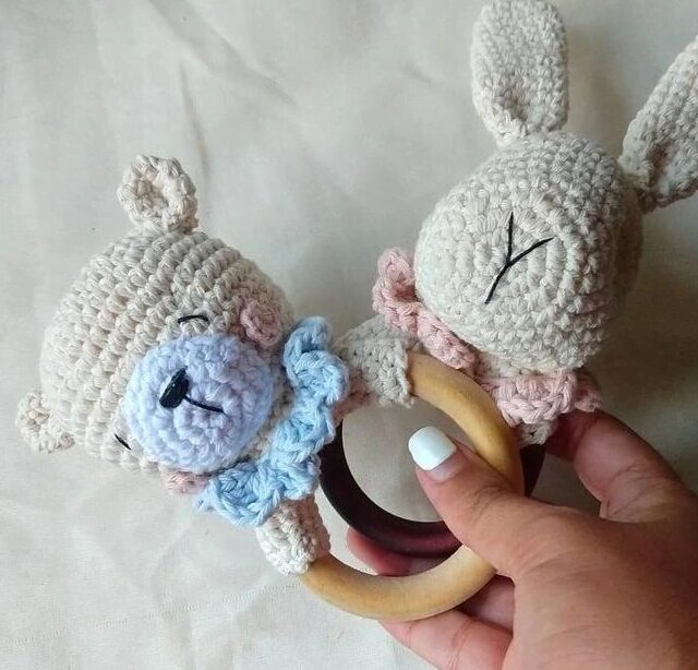 Un Primer Plano De Una Mano Que Sostiene Un Anillo De Dentición Para Bebé Tejido a Crochet Con Dos Animales El Anillo Fabricado En Madera Tiene Por Un Lado Un Osito Tejido a Crochet Con Un Lazo Azul Y Un Conejito Tejido a Crochet Por El Otro Lado Con Un Lazo Rosa El Fondo Es Una Tela Lisa De Color Claro