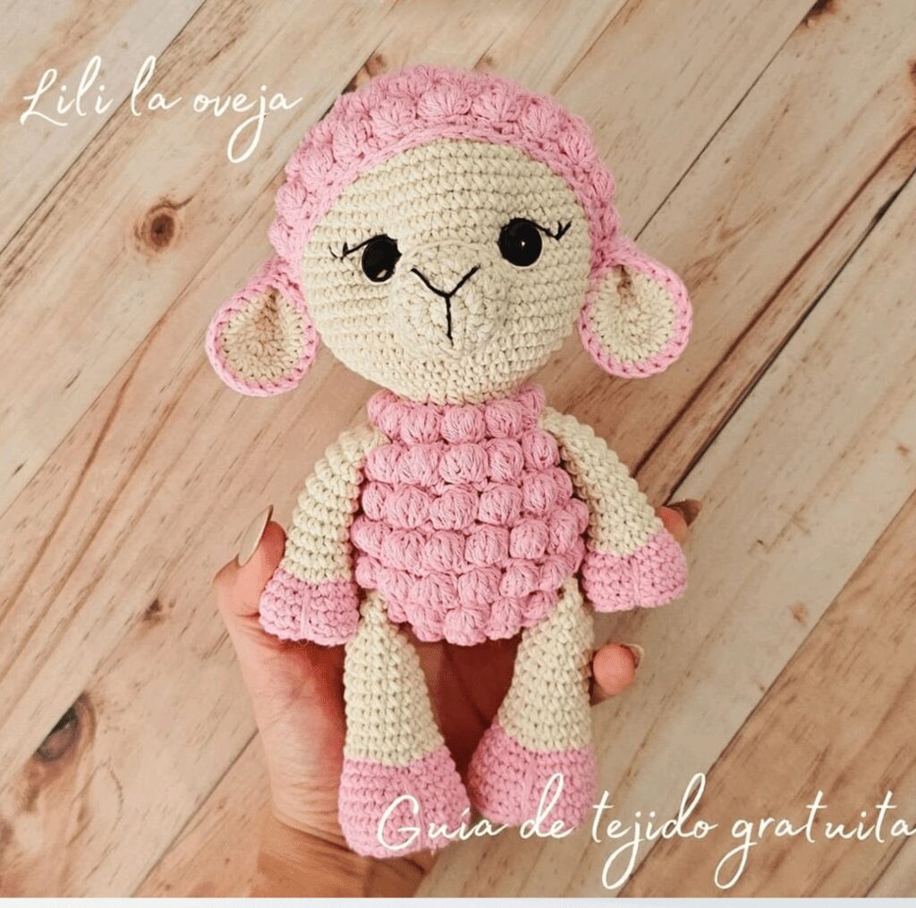 Una Mano Sostiene Un Juguete De Oveja De Ganchillo Con Colores Rosa Y Crema La Oveja Amigurumi Tiene Un Cuerpo Y Orejas De Color Rosa Texturizado Con Una Cara Sencilla Y Linda El Texto Dice lili La Oveja Y guía De Tejido Gratuita En La Imagen El Fondo Es Una Superficie De Madera