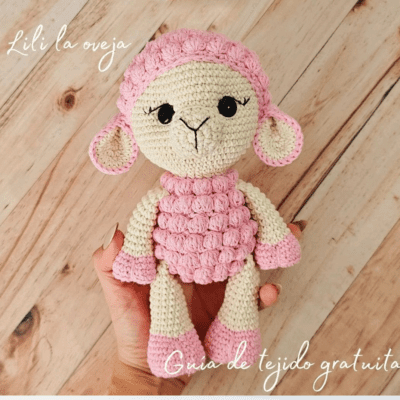 Una mano sostiene un juguete de oveja de ganchillo con colores rosa y crema. La oveja amigurumi tiene un cuerpo y orejas de color rosa texturizado con una cara sencilla y linda. El texto dice "Lili la oveja" y "Guía de tejido gratuita" en la imagen. El fondo es una superficie de madera.