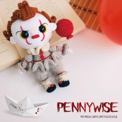 Un muñeco de crochet que se parece al personaje Pennywise de "It", y sostiene un globo rojo. El amigurumi de terror va vestido con un conjunto gris y rojo, con marcas rojas en la cara. Cerca hay un libro abierto y un barco de papel con la palabra "SS Georgie" escrita. El texto "PENNYWISE" está en la parte inferior.