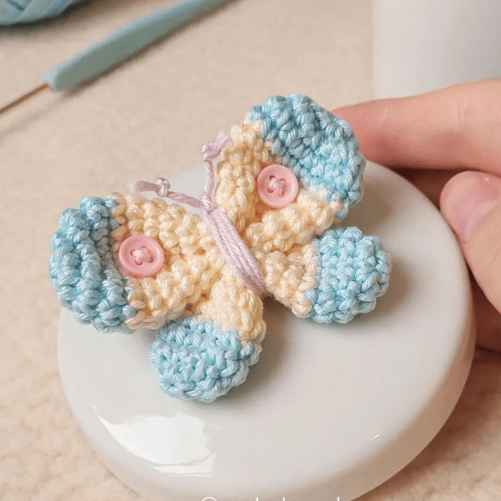 En Una Mano Se Sostiene Una Pequeña Mariposa Amigurumi Con Alas Color Crema Y Azul Decorada Con Botones Rosas La Mariposa Se Asienta Sobre Una Superficie Circular Blanca Al Fondo Se Puede Ver Un Ganchillo Y Un Hilo Azul Que Complementan Perfectamente El Intrincado Patrón