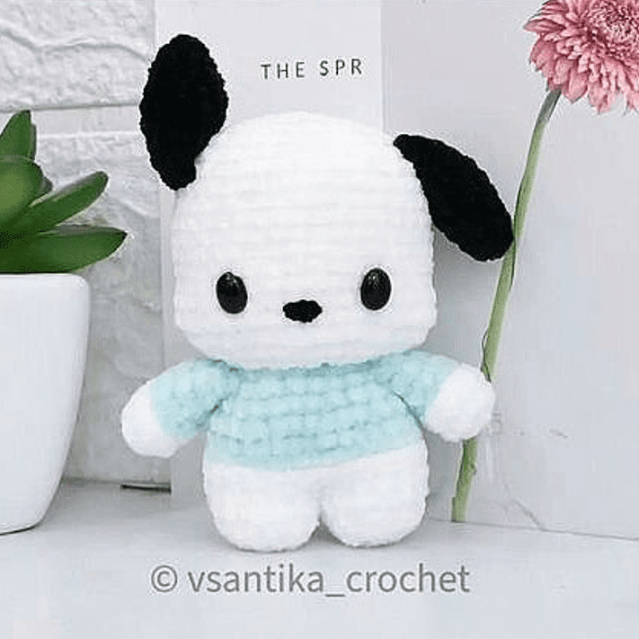 Un lindo animal de peluche de crochet blanco y negro con cabeza redonda, orejas negras y ojos negros lleva una camisa azul claro y blanca. Se encuentra junto a una planta en una maceta blanca y una planta alta y verde con una flor rosa sobre una superficie blanca. El texto en la parte superior dice "El SPR. Tejer un perrito.