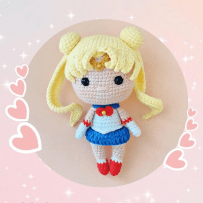 Una muñeca amigurumi Sailor Moon está centrada en la imagen, sobre un fondo rosa pastel adornado con destellos y gráficos de corazones. La muñeca tiene cabello rubio en sus característicos moños, un traje de marinero blanco y azul y botas rojas.