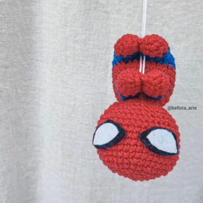 Una representación tejida a crochet de Spider-Man, colgando boca abajo de un hilo blanco de sus pies. La figura es predominantemente roja con los característicos ojos blancos y negros de Spider-Man. El fondo es una tela grisácea. El identificador de Instagram @bellota_arte es visible.