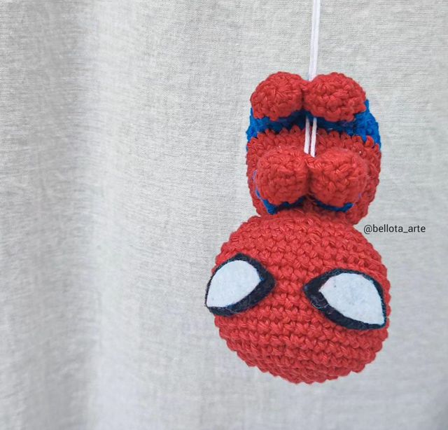 Una Representación Tejida a Crochet De Spider man Colgando Boca Abajo De Un Hilo Blanco De Sus Pies La Figura Es Predominantemente Roja Con Los Característicos Ojos Blancos Y Negros De Spider man El Fondo Es Una Tela Grisácea El Identificador De Instagram bellota arte Es Visible